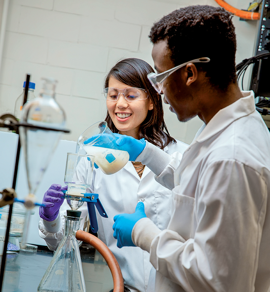 Lindsay Soh wears goggles, gloves, white lab coat in the lab with a student